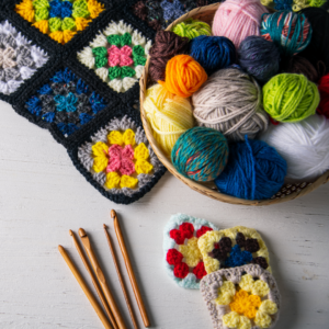 close up of crochet needles, yarn in bowl and colorful granny squares sewn together